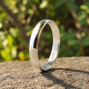 Puede incluir: Un anillo de banda de color plateado pulido con una superficie exterior lisa y redondeada. El interior del anillo presenta un grabado claro que dice 925, lo que indica la composición del metal. El anillo se muestra en posición vertical.