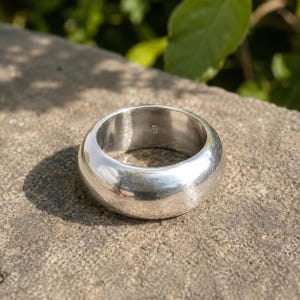 Peut inclure: Une large bague anneau en argent poli avec une surface extérieure lisse et arrondie. L'intérieur de la bague présente un poinçon 925 clair, indiquant la composition du métal. La bague possède une finition métallique réfléchissante sur tout son design.