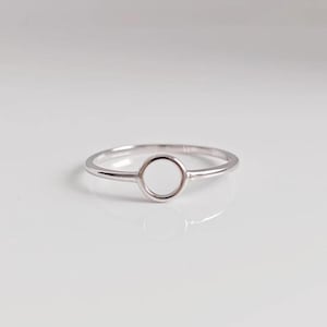 Puede incluir: Un anillo de tono plateado pulido que presenta una banda delgada y lisa que se conecta a un elemento circular abierto centrado. La superficie metálica es reflectante y de color uniforme, creando un diseño de bucle continuo.