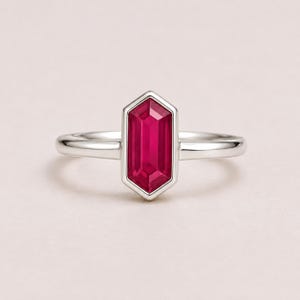 May include: A silver ring featuring a central, elongated hexagonal-cut vibrant red gemstone. The band is smooth and polished, holding the gemstone in a secure bezel setting. The design is symmetrical, showcasing the deep crimson color of the stone.