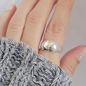 May include: A polished, smooth, silver-toned dome ring with a wide, rounded band. The metal surface is highly reflective, showing a clear sheen and a curved, solid shape that wraps around the finger. The ring has a simple, unadorned design.