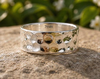 May include: A silver-toned ring featuring a hammered, dimpled texture across its outer surface. The interior of the band is clearly marked with the text 7mm, indicating the width of the metal band. The ring has a reflective, metallic finish.
