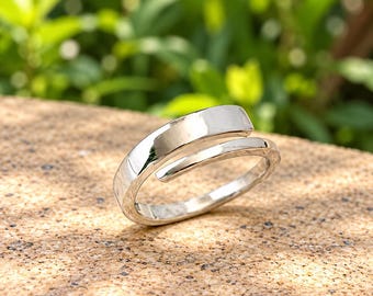 May include: A polished silver-tone bypass ring featuring a smooth, reflective surface and an open-ended design. The band curves around itself, creating a sleek, continuous loop with a metallic finish that catches the light.