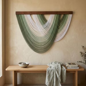 Op de afbeelding: Een wandhanger met meerdere strengen garen in saliegroen, olijfgroen en crèmetinten, opgehangen aan een horizontale donkerbruine houten balk. De garenstrengen zijn gerangschikt in overlappende, trapsgewijze U-vormige bogen.