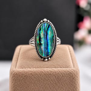Può includere: Un anello di forma ovale con un inserto in conchiglia di abalone lucida dalle tonalità iridescenti verdi, blu e viola. La conchiglia è incastonata in una montatura in metallo argentato con bordo a corda intrecciata e piccole sfere decorative.