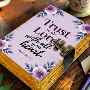 May include: A rectangular journal with a light purple cover featuring a floral pattern and the text Trust in the Lord with all your heart. Proverbs 3:5. The book has a dark metal latch closure and thick, textured, golden-yellow pages visible on the side.