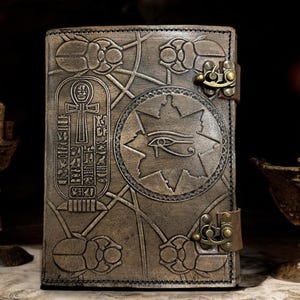 May include: A rectangular brown leather journal featuring embossed Egyptian motifs including scarab beetles, a central Eye of Horus within a star, and a cartouche with an ankh symbol. The cover is secured by two antique brass metal latches.