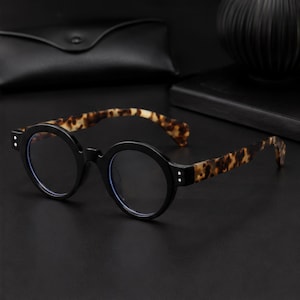 May include: Round spectacles featuring a solid black frame front with two small silver-tone rivets on each side. The temples display a brown and black tortoiseshell pattern. The lenses are clear with a subtle blue tint visible along the inner edges.