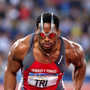 Peut inclure: Un coureur portant un débardeur de sport rouge, gris et noir avec le texte TRINIDAD & TOBAGO et le logo des anneaux olympiques. Le coureur porte des lunettes de soleil distinctives à monture argentée avec des verres rouges sur le front.