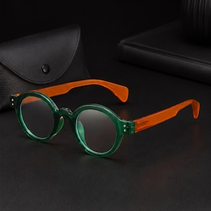 Könnte beinhalten: Runde Brille mit einem durchscheinenden smaragdgrünen Rahmen und leuchtend orangefarbenen Bügeln. Die Brille hat klare Gläser und kleine silberfarbene Nieten an den Scharnieren und wird mit einem schwarzen, strukturierten Etui geliefert.