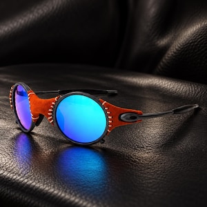 May include: Sunglasses with round, reflective blue lenses and a matte black frame. The bridge and outer edges of the lenses are wrapped in burnt orange suede leather, secured with visible white stitching. The temples are thin, black, and metallic.