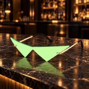 May include: Rimless sunglasses featuring sharp, triangular lime green lenses. The eyewear has thin, polished gold-tone metal temples that connect to the lenses with small screws, and the temple tips are finished with black plastic covers.