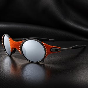 May include: Round sunglasses featuring reflective silver lenses and a black frame. The bridge and outer edges of the circular lenses are covered in textured orange suede material, secured with visible cream-coloured stitching along the perimeter.