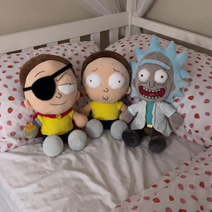 Può includere: Tre peluche raffiguranti personaggi di Rick e Morty. Un personaggio indossa una benda sull'occhio e un mantello rosso, un altro ha un'espressione preoccupata con una maglietta gialla, il terzo ha capelli blu a punta e un camice bianco.