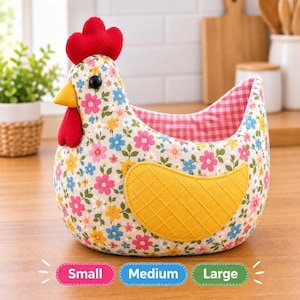 Puede incluir: Una cesta de tela con forma de gallina que presenta un exterior de color crema con un colorido estampado floral, una cresta y barbillas de color rojo brillante, un pico amarillo y un ala acolchada amarilla. El interior está forrado con tela de cuadros rosas y blancos.