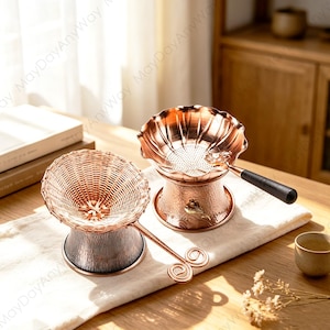 May include: Two copper-coloured tea strainers with stands. One features a woven metal mesh top and a handle with a double-spiral end. The other has a scalloped edge, a perforated base, and a black handle with the word Grace printed on it.
