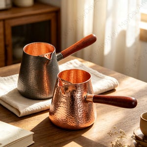 Peut inclure: Deux cafetières en métal martelé avec des manches en bois. L'une présente un extérieur gris foncé avec un intérieur en cuivre, tandis que l'autre est entièrement cuivrée. Les deux possèdent un bec verseur avec filtre et un long manche en bois.