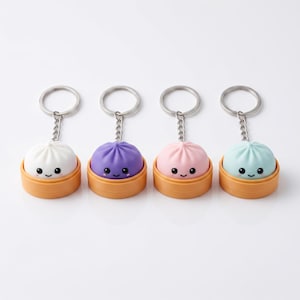 Peut inclure: Quatre porte-clés représentant des figurines de brioches vapeur miniatures en blanc, violet, rose et bleu clair. Chaque brioche a un visage souriant avec des yeux noirs et des joues roses, posée dans une base orange en forme de panier vapeur.