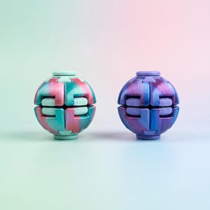 Op de afbeelding: Twee bolvormige fidget-speeltjes met gesegmenteerde, in elkaar grijpende panelen. De linkerbol heeft een gemarmerde mix van mintgroen en roze, terwijl de rechterbol een gemarmerde combinatie van dieppaars en blauw toont, beide met een naad.