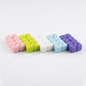 Op de afbeelding: Een set van vijf rechthoekige fidget-speeltjes op een rij, elk bestaande uit een schakelketting van kleine blokjes met een textuur van honingraatpatronen. De kleuren zijn lichtroze, limoengroen, wit, lichtblauw en paars.