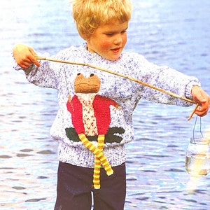 May include: A child's blue and white knit sweater with a frog design. The frog has a brown head, red jacket, and yellow and white striped legs. The child is holding a fishing rod and a glass jar.