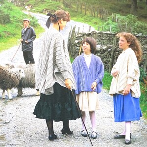 May include: A rural scene with people and sheep on a gravel path. A woman in a gray sweater and black skirt stands with two girls, one in a blue cardigan and the other in a white sweater and blue skirt. A man in a raincoat herds sheep with a stick.