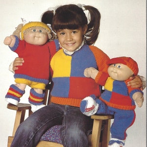 May include: A young girl sits holding two Cabbage Patch Kids dolls. The dolls and the girl wear color-blocked knit outfits in red, blue, yellow, and orange. The dolls have blonde and red hair, and the girl has dark hair.