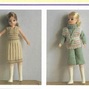 May include: Two dolls wearing knitted outfits. One doll is in a beige sleeveless top and skirt with white boots. The other doll is in a green turtleneck, vest, and pants with white boots. The vest has a pink and white pattern.
