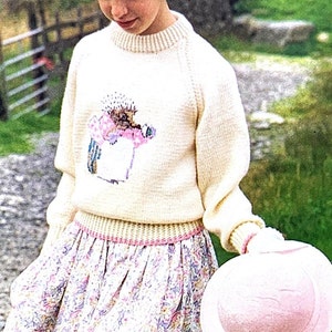 May include: A cream-coloured sweater with a floral design, paired with a floral print skirt and a pink hat. The sweater features a crew neck and long sleeves. The skirt has a pink waistband. The hat is light pink.