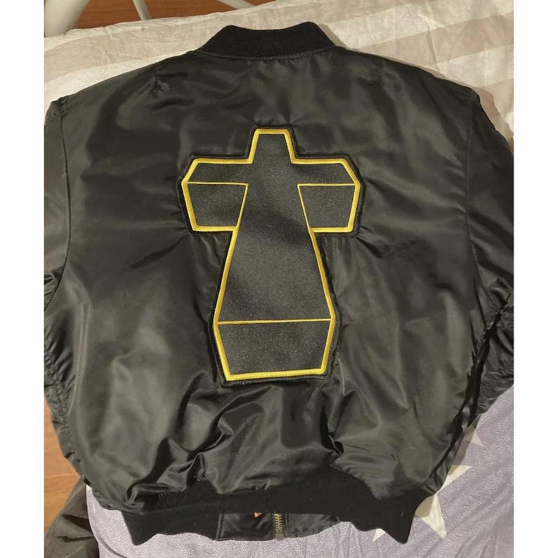 Close-up of a Justice Cross satin varsity jacket highlighting the embroidered cross design, smooth satin fabric, detailed stitching, ribbed trim, and classic bomber construction. This vintage-inspired unisex streetwear jacket offers a distinctive retro look and can be paired with denim, sneakers, casual trousers, or layered clothing.