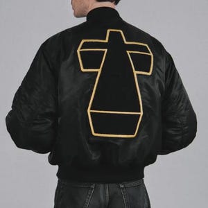 Puede incluir: Una chaqueta bomber de satén negro con un gran emblema de cruz centrado en la espalda. El emblema está hecho de tela negra con un borde grueso bordado en color dorado. La chaqueta tiene un cuello acanalado y mangas largas de acabado liso.
