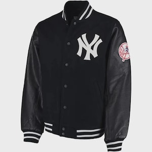 Op de afbeelding: Een zwart varsity-jack met een wit New York Yankees-logo op de borst en een ronde team-patch op de mouw. Het jack heeft zwarte leren mouwen, een sluiting met drukknopen aan de voorzijde en wit gestreepte boorden bij de kraag en mouwen.