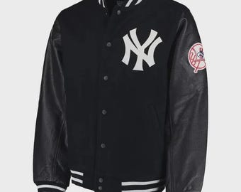 Puede incluir: Una chaqueta universitaria negra con el logotipo blanco de los New York Yankees en el pecho y un parche circular del equipo en la manga. La chaqueta tiene mangas de cuero negro, cierre frontal con botones a presión y ribetes a rayas blancas.