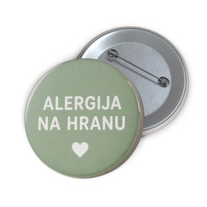 May include: A round pinback button with a sage green background featuring the white text ALERGIJA NA HRANU and a small white heart icon below the text. The back of the button is metallic silver with a standard pin closure mechanism attached.