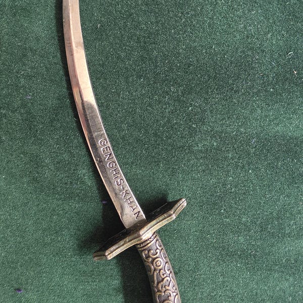 Könnte beinhalten: Eine metallene, geschwungene Klinge mit der Gravur GENGHIS-KHAN auf der Oberfläche. Der Griff weist ein komplexes Prägemuster auf, wobei ein rechteckiger Parierstange den Griff von der Klinge trennt, alles in einem bronzefarbenen Ton.