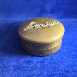Könnte beinhalten: Ein runder Messingbehälter mit einem Deckel, der den geprägten Schriftzug "Sunkist" trägt. Die Metalloberfläche hat einen gedämpften, gealterten Goldton mit sichtbarer Patina und einer glatten Oberfläche auf der Oberseite und den Seiten.
