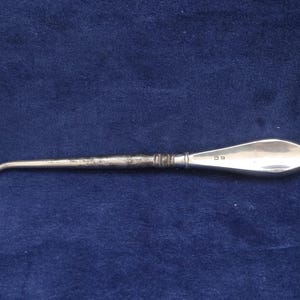 May include: A silver button hook with a polished, teardrop-shaped handle and a slender, textured metal shaft. The handle features small, stamped hallmarks near the transition to the shaft, which ends in a curved hook for fastening buttons.