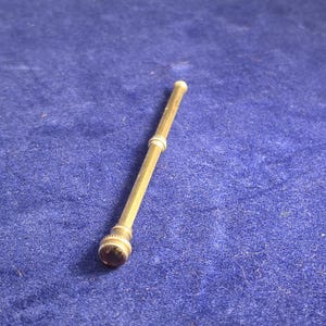 Puede incluir: Una herramienta cilíndrica y delgada de metal dorado que presenta una superficie texturizada con un patrón de rayas cruzadas a lo largo de su longitud. El objeto incluye una pequeña incrustación redondeada de piedra o vidrio oscuro en un extremo y una banda decorativa estriada cerca del centro.