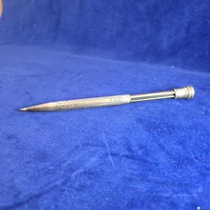 Peut inclure: Un porte-mine métallique doté d'un corps cylindrique fin avec un motif en chevrons gravé le long du fût. L'instrument d'écriture présente une pointe effilée et un capuchon arrondi et strié à l'extrémité opposée, avec une finition polie.