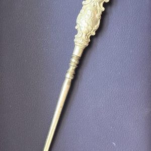 May include: A gold-toned metal button hook featuring an ornate, embossed handle with detailed scrollwork and cherub motifs. The long, slender shaft transitions into a curved hook at the end, designed for fastening buttons on garments or footwear.