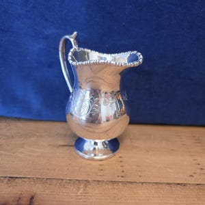 May include: A small silver-toned creamer pitcher featuring a polished metallic finish, a curved handle, a scalloped rim with beaded detailing, and an intricate floral engraving on the rounded body, supported by a circular pedestal base.