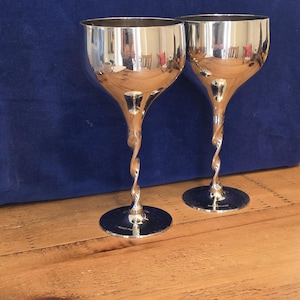 May include: Two silver-plated goblets with smooth, rounded bowls and twisted, spiral stems. Each goblet features a circular base and a polished, reflective metallic surface that shows distorted reflections of the surrounding environment.