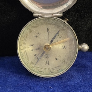 May include: A vintage silver-toned metal pocket compass with a hinged lid. The circular face features a weathered off-white dial with black cardinal directions, degree markings, and the text "MADE IN FRANCE" printed clearly below the centre.