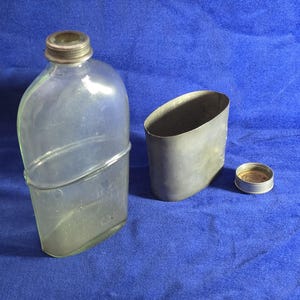 Peut inclure: Une gourde en verre transparent avec un goulot en métal fileté, accompagnée d'un étui en métal de forme ovale et d'un bouchon à vis circulaire assorti, présentant une doublure en liège visible sur sa face intérieure.