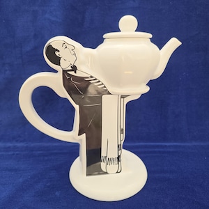 Op de afbeelding: Een witte keramische theepot in de vorm van een butler in een zwart smoking en vlinderdas. De figuur houdt een wit theepotlichaam met deksel op een dienblad, met een witte handdoek over de arm gedrapeerd. Het handvat is de rug van de butler.