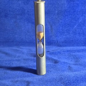 May include: A cylindrical metal hourglass with a matte silver finish and a small knob at the top. A vertical cutout in the centre of the tube reveals a glass bulb containing light brown sand, showing the flow of time through the narrow neck.