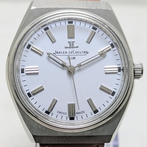 Może przedstawiać: Zegarek na rękę Jaeger-LeCoultre Club ze stali nierdzewnej z białą tarczą, srebrnymi indeksami w kształcie pałeczek i brązowym skórzanym paskiem. Tarcza zawiera logo Jaeger-LeCoultre, napis CLUB oraz słowa SWISS MADE na dole.