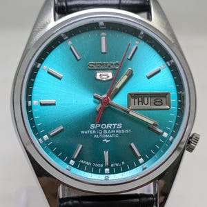 May include: A Seiko 5 Sports automatic wristwatch with a sunburst teal dial, silver-tone hour markers, and a day-date window at the three o'clock position. The watch features a red second hand, a stainless steel case, and a black leather strap.