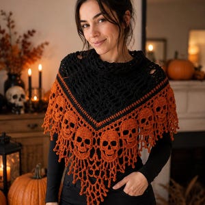 Puede incluir: Un chal triangular de ganchillo con una sección superior negra de patrón calado y un borde inferior de hilo naranja vibrante. La sección naranja está decorada con un patrón repetitivo de motivos de calaveras tejidas y flecos colgantes.
