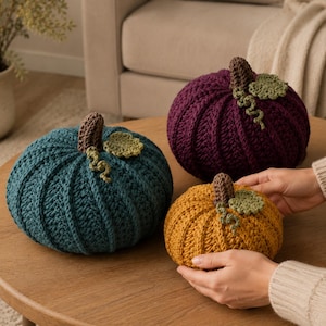 May include: Three crocheted pumpkins in teal, deep purple, and mustard yellow. Each pumpkin features a brown stem, a small green leaf, and a curly vine. The pumpkins have a ribbed texture created by the crochet stitches and are round in shape.